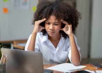 Frau sitzt am Schreibtisch und schaut angestrengt auf den Laptop, während sie sich mit den Händen die Schläfen hält