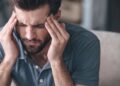 Mann in blauem Poloshirt massiert mit den Fingerspitzen beide Schläfen und verzieht das Gesicht vor Schmerzen