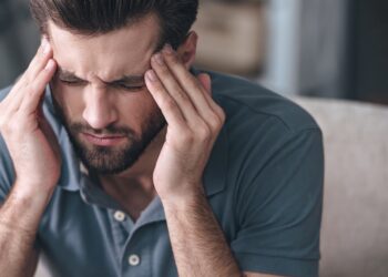 Mann in blauem Poloshirt massiert mit den Fingerspitzen beide Schläfen und verzieht das Gesicht vor Schmerzen