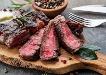 Medium rare Steak aufgenschitten auf einem Holzbrett mit Pfefferkörnern, Messer und Gabel daneben