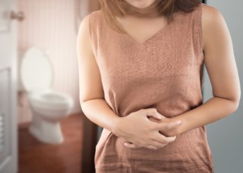 Eine Frau steht vor der geöffneten Tür zur Toilette und hält sich die Hände an den schmerzenden Bauch.