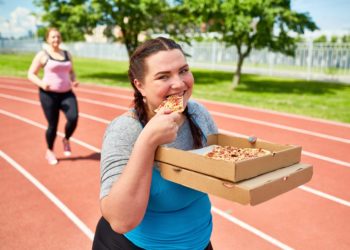 Zwei Frauen joggen über eine Laufstrecke und essen dabei Pizza.