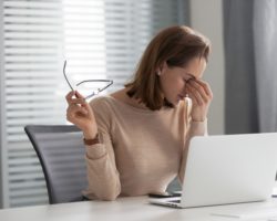 Erschöpfte junge Frau sitzt vor ihrem Laptop und fasst sich an ihre Nasenwurzel