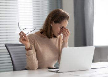 Erschöpfte junge Frau sitzt vor ihrem Laptop und fasst sich an ihre Nasenwurzel