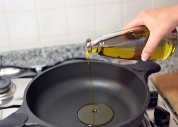 Aus einer durchsichtigen Flasche wird Speieseöl in eine Pfanne auf einem Herd geschüttet