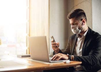 Mann mit Mundschutz sitzt vor einem Laptop und hält sein Smartphone in der rechten Hand