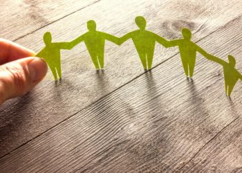 Menschen aus grünem Papier halten sich an den Händen, am linken Ende fassen Daumen und Zeigefinger einer echten linken Hand die Hand einer Papierfigur