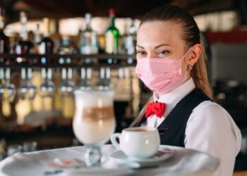 Eine weibliche Bedienung mit Mund-Nasen-Schutz serviert Kaffee