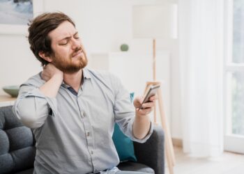 Mann mit schmerzverzerrtem Gesicht fasst sich an den Nacken und blickt auf sein Smartphone