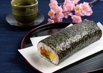 Eine Sushi-Rolle auf einem Teller vor einer Tasse mit Tee und Blumen auf einem Tisch