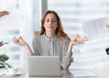 Junge Frau sitzt vor einem Laptop und macht eine Meditationsübung, während von allen Seiten Aufgaben an sie herangetragen werden