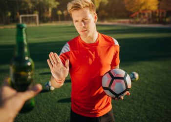 Ein Fußballer mit eine Fußball in der Hand weist eine angebotene Flasche Bier zurück