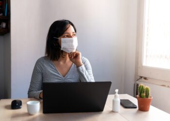 Frau mit Mund-Nasen-Bedeckung sitzt vor ihrem Laptop und sieht aus dem Fenster