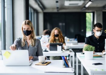 Mehrere Personen sitzen mit Mund-Nasen-Bedeckung vor ihrem Computer in einem Büro