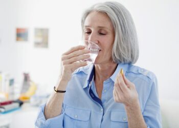 Ältere Frau trinkt aus einem Glas Wasser und hält eine Omega-3-Kapsel