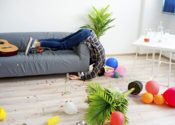 Ein Mann schläft halb aus dem Sofa hängend in einem unordentlichen Zimmer.