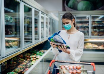 Frau mit OP-Maske liest Beschreibung eines Tiefkühlprodukts im Supermarkt