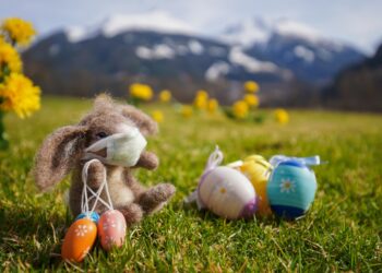 Ein Osterhase mit Mundschutz sitzt auf einer Wiese neben drei Ostereiern.