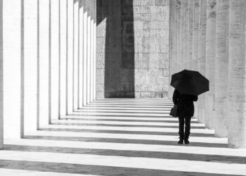 Mann in Schwarz gekleidet mit schwarzem Regenschirm geht einen Gang lang.