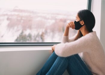 Eine Frau mit Mundschutz sitz vor einem Fenster und schaut nach draußen.