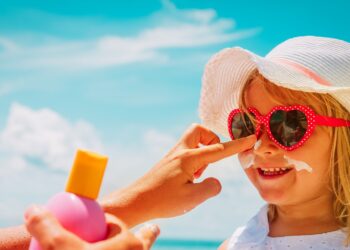 Frau cremt Mädchen mit Sonnenhut und Sonnenbrille das Gesicht mit Sonnenschutzcreme ein.