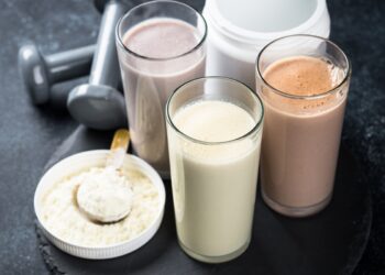 Verschiedene Proteinshakes in Gläsern, neben einem Eiweißpulver und Hanteln