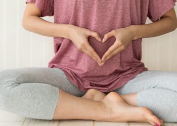 Eine Person sitzt im Schneidersitz auf den Boden und formt mit beiden Händen ein Herz vor dem Bauch.