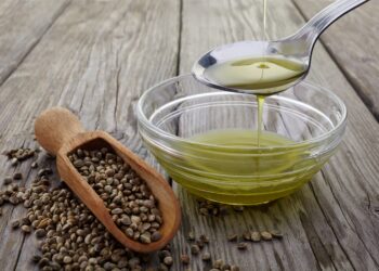 Pouring hemp oil into glass bowl and hemp seeds on wood