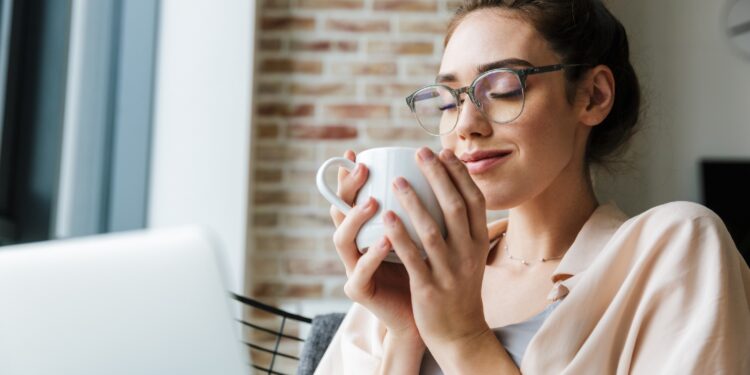 Frau sitzt auf dem Sofa und hält mit beiden Händen eine Tasse Kaffee.