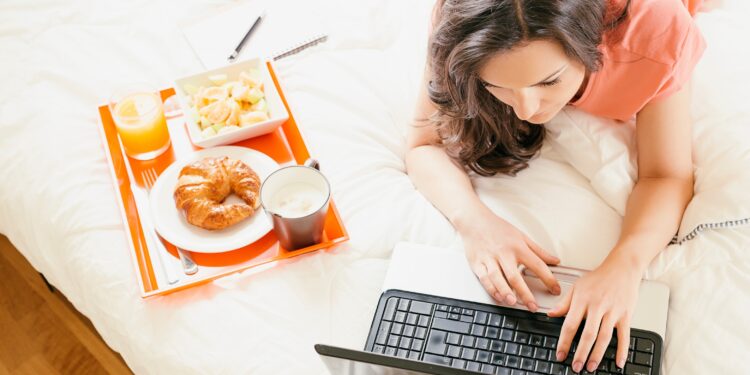Frau liegt auf dem Bauch im Bett und tippt auf dem Laptop neben ihr steht ein Tablett mit frischem Orangensaft Croissant und Kaffee